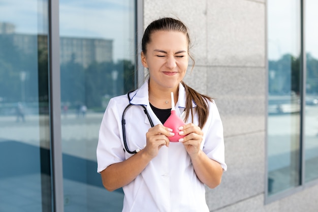 Medicina, assistenza sanitaria, professione e concetto di persone - Una divertente dottoressa europea in camice bianco con uno stetoscopio soffia aria su se stessa da un clistere che tiene tra le mani all'esterno