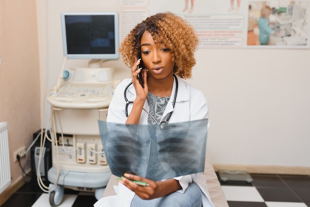 Medicina, chirurgia e concetto di sanità - medico o chirurgo femminile afroamericano in camice bianco che esamina i raggi x sopra il personale dell'ospedale su priorità bassa