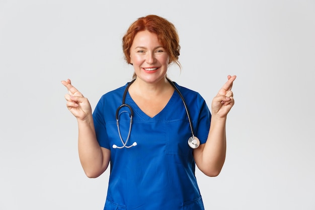 Medicina, concetto di assistenza sanitaria. Operaio medico femminile ottimista che spera in meglio. Redhead medico in blu frega sorridendo e incrocia le dita, buona fortuna, esprimendo il desiderio