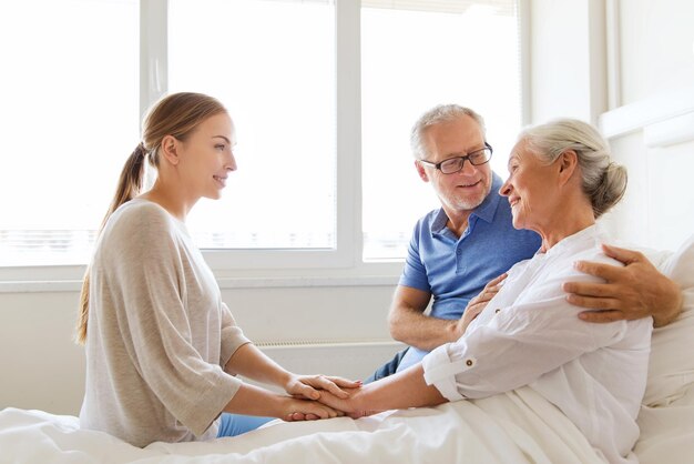 medicina, supporto, assistenza sanitaria familiare e concetto di persone - uomo anziano felice e giovane donna che visitano e acclamano sua nonna sdraiata a letto nel reparto ospedaliero