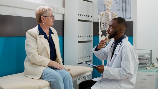 Medico afroamericano che consulta la donna anziana nell'ufficio del medico, spiegando la diagnosi medica all'appuntamento della visita di controllo. Medico e paziente che parlano del supporto sanitario.