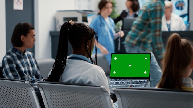 Medico afroamericano che lavora con lo schermo verde sul computer portatile, seduto nella hall della reception dell'ospedale. Medico che utilizza sfondo chroma key isolato con copyspace vuoto e modello mockup.