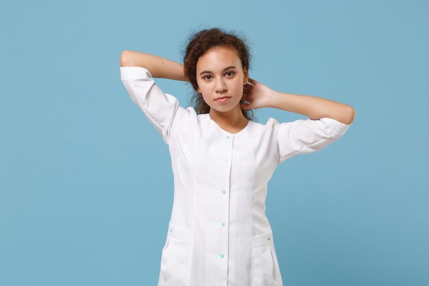 Medico afroamericano donna isolata su sfondo blu in studio. Medico femminile in abito medico bianco che si tiene per mano dietro la testa. Personale sanitario, salute, concetto di medicina. Simula lo spazio della copia.