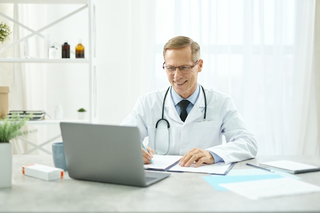 Medico allegro che utilizza laptop e compila la cartella clinica del paziente