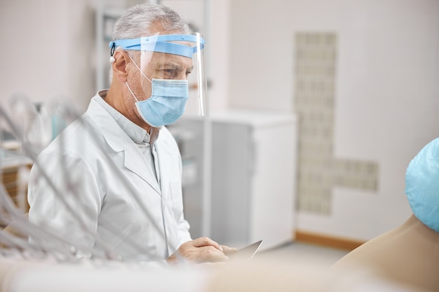 Medico anziano che indossa un camice da laboratorio e una visiera con in mano un tablet mentre è in clinica