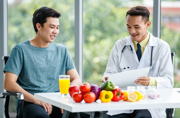 Medico asiatico positivo che sorride e mostra il tavolo al paziente di sesso maschile mentre è seduto alla scrivania con cibo sano in clinica