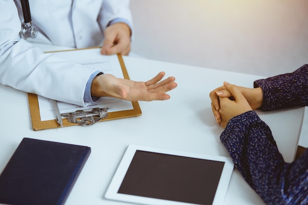 Medico barbuto sconosciuto e donna paziente che discutono dell'attuale esame sanitario mentre si siedono in una clinica soleggiata, primo piano.