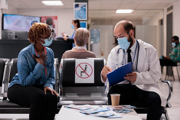 Medico che consulta un paziente afroamericano nella hall della clinica, situato nell'area di attesa della reception dell'ospedale per partecipare all'appuntamento di controllo. Fare un esame sanitario durante la pandemia di covid 19.