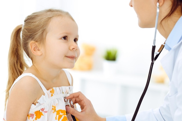 Medico che esamina un bambino con lo stetoscopio in clinica soleggiata. La paziente sorridente felice della ragazza vestita con un vestito di colore brillante è alla normale ispezione medica.