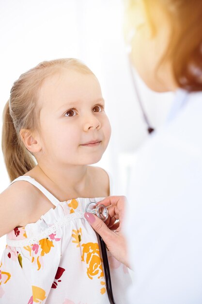 Medico che esamina un bambino con lo stetoscopio in clinica soleggiata. La paziente sorridente felice della ragazza vestita con un vestito di colore brillante è alla normale ispezione medica.