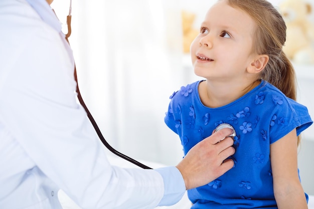 Medico che esamina un bambino con lo stetoscopio in clinica soleggiata. La paziente sorridente felice della ragazza vestita in vestito blu è alla normale ispezione medica.