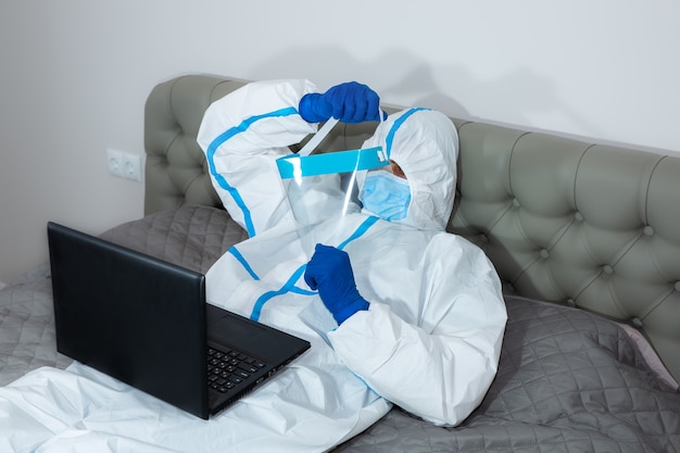 Medico che indossa tuta protettiva medica, occhiali, maschera e guanti che lavorano sul laptop, sdraiato sul letto a casa. Protezione dall'epidemia di virus. Coronavirus (COVID-19). concetto di assistenza sanitaria.