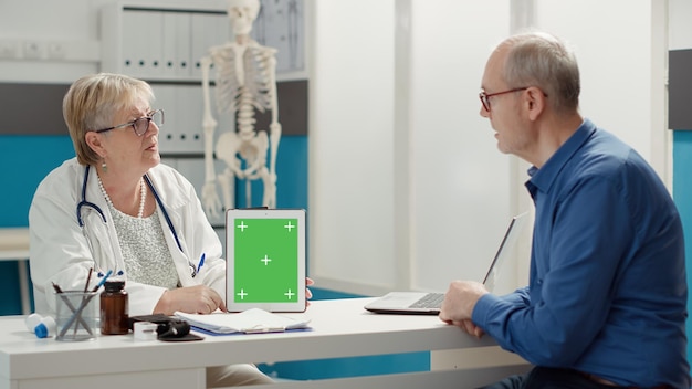 Medico che mostra tablet con schermo verde al paziente in pensione all'appuntamento per l'esame nel gabinetto dell'ospedale. Modello di chiave cromatica isolato con copyspace vuoto e sfondo mockup su gadget digitale.