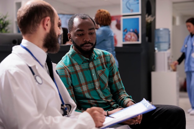 Medico che parla con un paziente maschio in sala d'attesa, utilizzando il rapporto di controllo medico per fornire diagnosi e trattamento della malattia. Medico e uomo che fanno consulenza sanitaria in clinica.