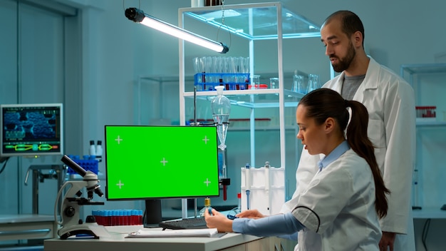Medico che parla con un tecnico di laboratorio e lavora con un computer desktop con schermo mock-up verde. Ricercatore di laboratorio uomo che discute con il medico sullo sviluppo del vaccino guardando i campioni di sangue