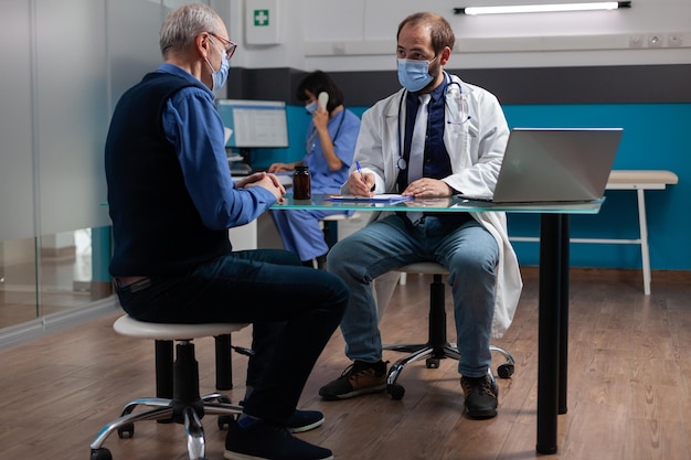 Medico che prende appunti sugli appunti e parla con il pensionato durante la consultazione durante la pandemia di covid 19. Medico che fa visita di controllo con il paziente, scrivendo la diagnosi sul file di rapporto in ufficio.