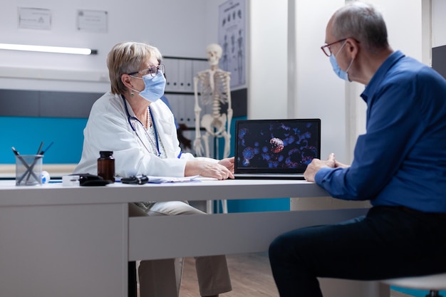 Medico che spiega i batteri del coronavirus sul laptop a un uomo anziano, che indossa una maschera facciale in studio medico. Medico donna che mostra una rappresentazione visiva del virus covid 19 durante la visita di controllo con il paziente