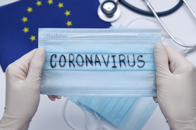 Medico che tiene la maschera con la parola Coronavirus sopra gli articoli medici e il primo piano della bandiera dell'Unione europea