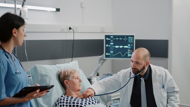 Medico che utilizza lo stetoscopio per la misurazione del battito cardiaco sul paziente malato nel letto del reparto ospedaliero. Medico che fa consulenza sanitaria con una donna anziana mentre l'infermiera con tablet dà assistenza