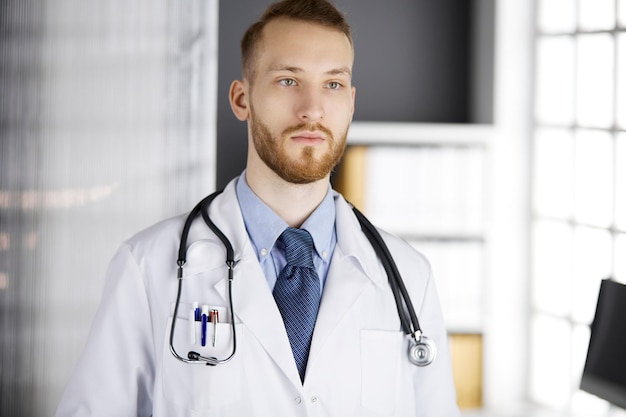 Medico con la barba rossa in piedi dritto in clinica vicino al suo posto di lavoro. Ritratto di medico. Concetto di medicina e assistenza sanitaria.