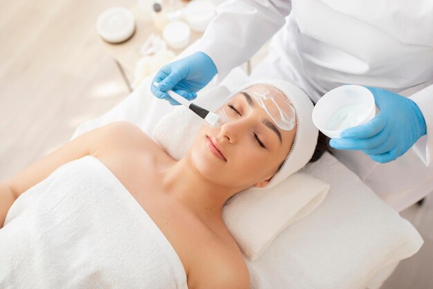 Medico cosmetologo professionista che applica maschera idratante sul viso di una bella donna indiana