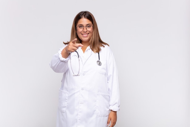 Medico della giovane donna che indica alla macchina fotografica con un sorriso soddisfatto, sicuro, amichevole, scegliendo voi