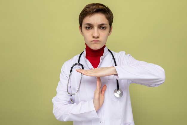 Medico della ragazza in camice bianco con lo stetoscopio intorno al collo con la faccia seria che fa il gesto di time out con le mani che stanno sul green