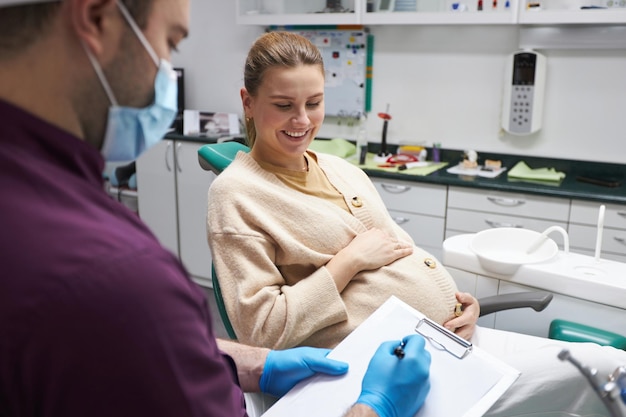 Medico dentista che prescrive un trattamento a una donna incinta che sorride mentre si siede sulla poltrona del dentista