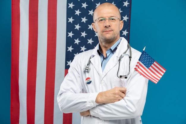 Medico di mezza età sicuro in occhiali e cappotto bianco che tiene piccola bandiera americana mentre levandosi in piedi contro stelle e strisce