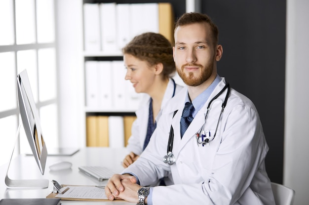 Medico di sesso maschile con un collega che discute la terapia attuale della malattia mentre è seduto al posto di lavoro in clinica. Lavoro di squadra in medicina.