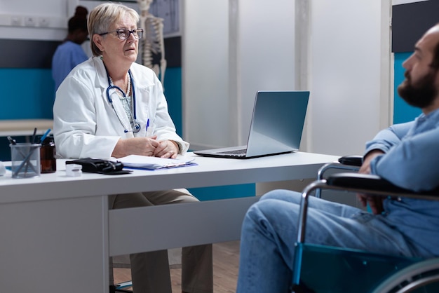 Medico donna che discute con l'uomo seduto in sedia a rotelle alla visita di controllo. Medico presente all'appuntamento di consultazione per il paziente con disabilità. Persone che parlano nel gabinetto