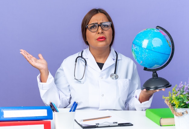 Medico donna di mezza età in camice bianco con stetoscopio con gli occhiali che tiene il globo guardando davanti con il viso serio accigliato con il braccio fuori seduto al tavolo sul muro blu