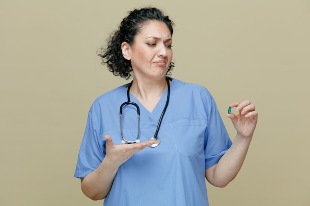 Medico donna di mezza età scontento che indossa uniforme e stetoscopio intorno al collo che mostra la capsula che lo guarda mostrando la mano vuota isolata su sfondo verde oliva