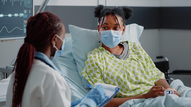 Medico e paziente che discutono del trattamento contro la malattia indossando maschere per il viso. Medico di medicina generale che consulta una donna malata con ossimetro, collegato al monitor della frequenza cardiaca in reparto ospedaliero.