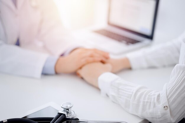 Medico e paziente seduti uno di fronte all'altro al tavolo nell'ufficio della clinica. L'attenzione si concentra sulle mani del medico femminile che rassicurano la donna, solo le mani, da vicino. Concetto di medicina.