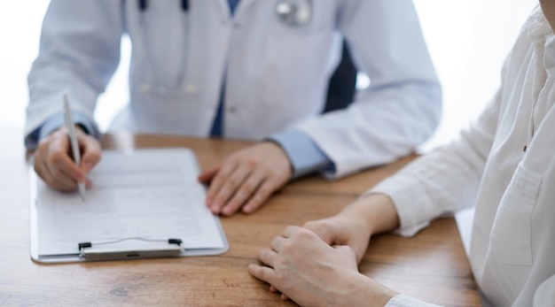 Medico e paziente seduto alla scrivania in legno in clinica. L'attenzione si concentra sulle mani del medico femminile che riempiono un modulo di registrazione. Concetto di medicina