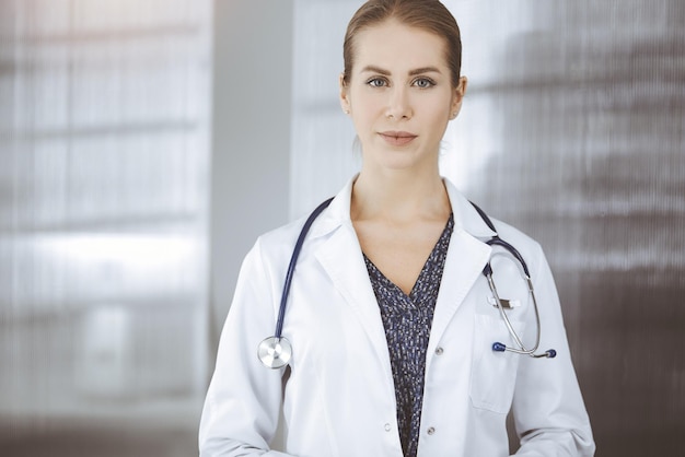 Medico femminile allegro che sta nella clinica soleggiata. Ritratto di donna-medico sorridente amichevole. Concetto di medicina.