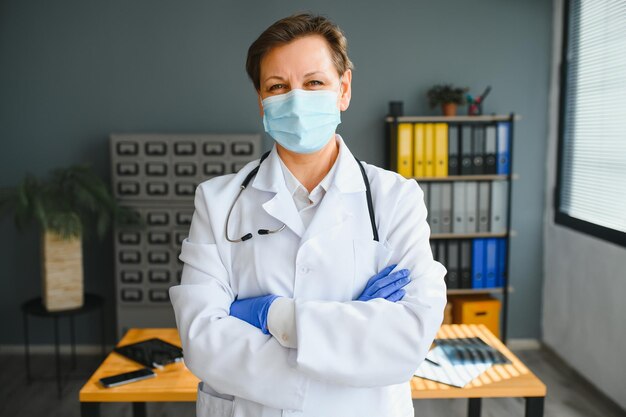 Medico femminile anziano che indossa maschera facciale e camice medico bianco in piedi in ospedale Ritratto