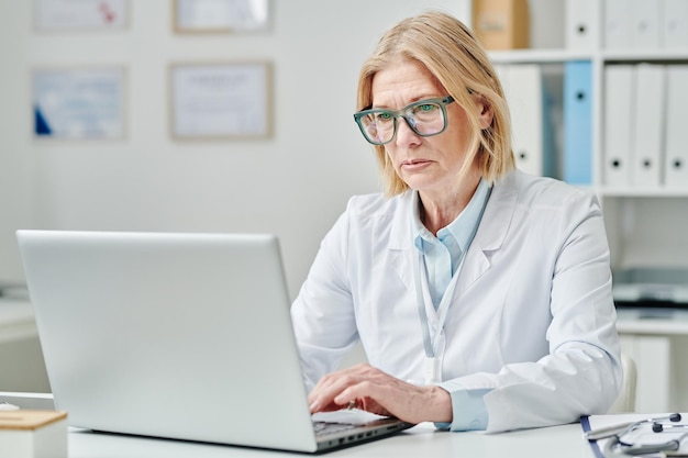 Medico femminile biondo maturo serio che digita sulla tastiera del computer portatile in ufficio