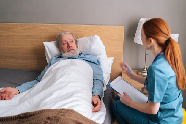 Medico femminile che annota diagnosi e sintomi nella storia medica negli appunti, parlando con un paziente maschio anziano malato sdraiato sul letto a casa. Uomo anziano che riceve un consulto medico in ospedale.