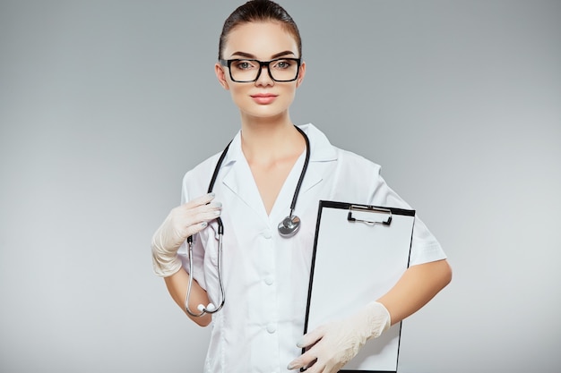 Medico femminile con capelli castani e trucco nudo indossando l'uniforme medica bianca, occhiali, stetoscopi e guanti bianchi su sfondo grigio studio e note di partecipazione.