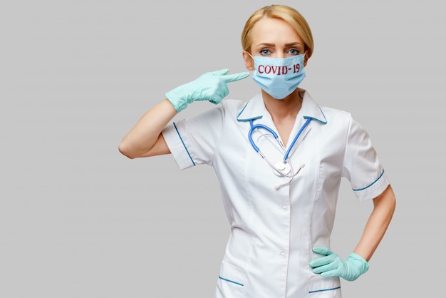 Medico femminile con lo stetoscopio che indossa i guanti protettivi del lattice e della maschera sopra fondo grigio chiaro