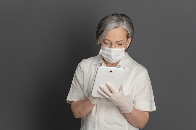 Medico femminile dai capelli grigi che lavora con il blocco note. Donna caucasica che indossa uniforme protettiva bianca, maschera e occhiali utilizzando la tavoletta digitale. Isolato su grigio indietro con copyspace a sinistra