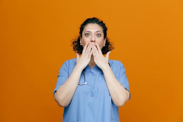 medico femminile di mezza età sorpreso che indossa uniforme e stetoscopio intorno al collo tenendo le mani sulla bocca guardando la fotocamera isolata su sfondo arancione