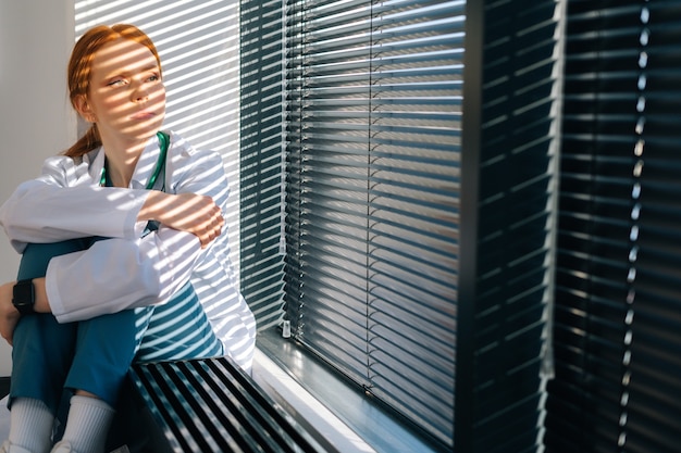 Medico femminile frustrato infelice che piange seduto vicino alla finestra in una giornata di sole. Il medico stressato della giovane donna turbata si sente preoccupato per la negligenza professionale. .