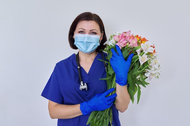 Medico femminile in maschera medica uniforme blu con bouquet di fiori su sfondo chiaro