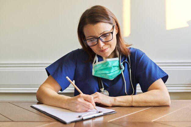 Medico femminile in uniforme blu con stetoscopio, maschera medica protettiva seduta al tavolo e scrivendo negli appunti. Ritratto del primo piano del medico sicuro professionale