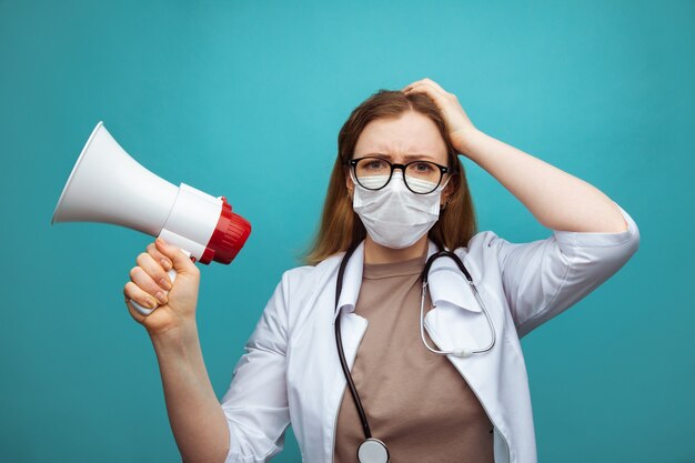 Medico femminile perplesso nella maschera di protezione dell'abito medico isolata sull'azzurro