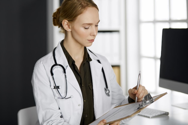 Medico femminile sorridente allegro che utilizza appunti in clinica. Ritratto di donna medico amichevole sul posto di lavoro. Servizio medico perfetto in ospedale. Concetto di medicina.