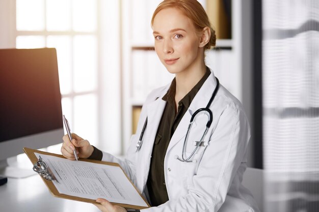 Medico femminile sorridente allegro che utilizza appunti nella clinica soleggiata. Ritratto di donna medico amichevole sul posto di lavoro. Servizio medico perfetto in ospedale. Concetto di medicina.